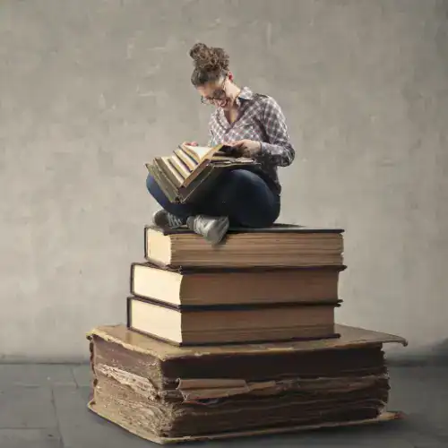 Woman sitting on top of a stack of oversized books, reading a small book, symbolizing knowledge and learning, with a plain background.